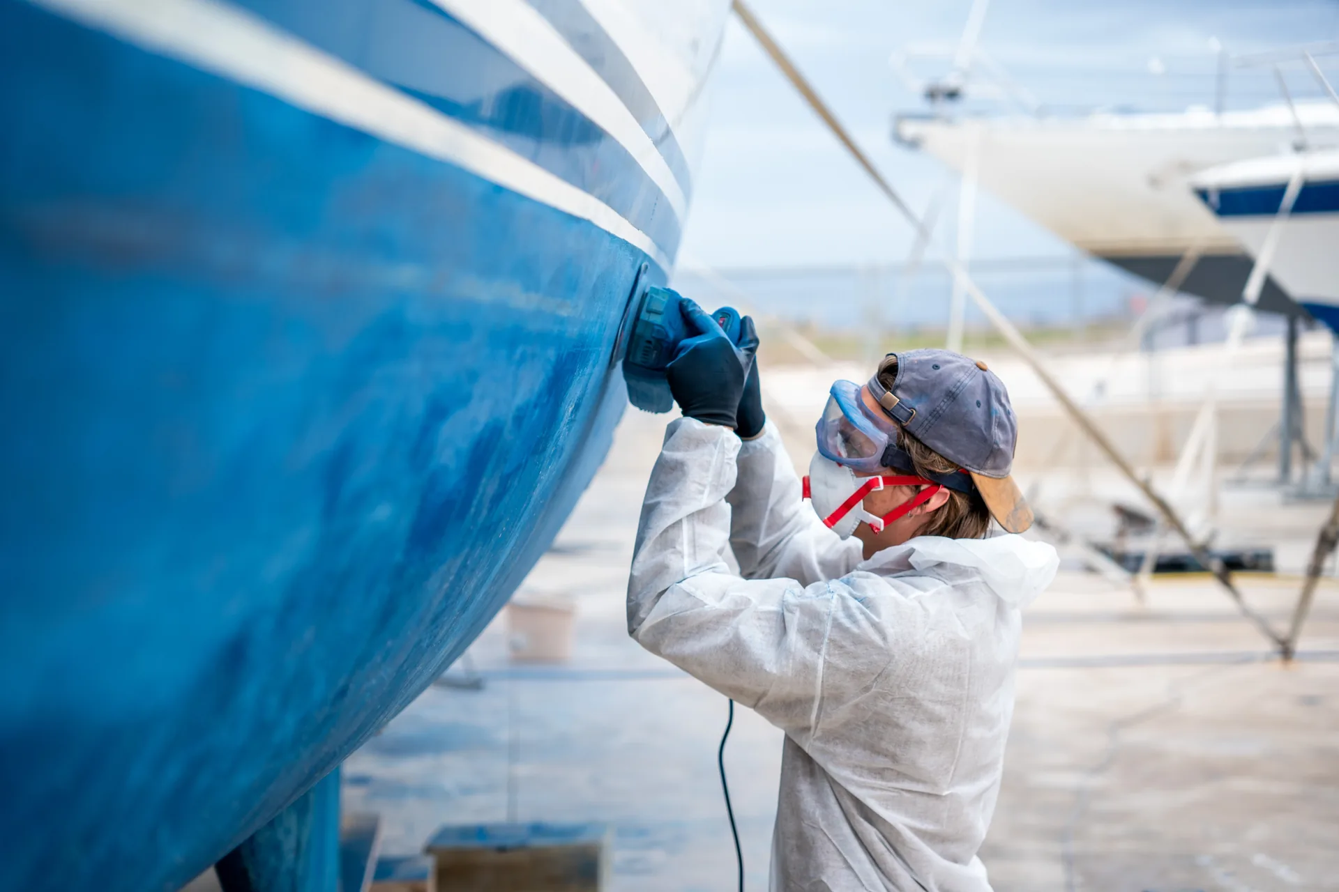 Worker in protective clothing sanding a large blue boat hull in a boatyard